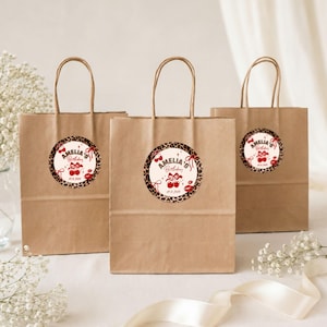 May include: Three brown paper gift bags with handles, each featuring a circular sticker with a leopard print border. The sticker displays the text "AMELIA'S Birthday" in red, accompanied by illustrations of red cherries, bows, and lip prints.