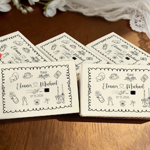 May include: Off-white matchboxes with black decorative borders and illustrations. The boxes are personalized with the names "Eleanor & Michael" and the date "07.15.2026". A few matches with red tips are visible.