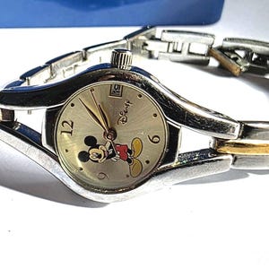 May include: Silver and gold-toned Disney watch with a Mickey Mouse graphic on the face. The watch has a silver band with gold accents and a date display at the 3 o'clock position. The watch is approximately 7 inches in length.