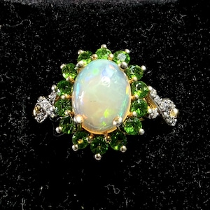 May include: A gold ring featuring a large, oval opal gemstone surrounded by small, vibrant green gemstones. The opal has a milky white appearance with iridescent flashes. The ring's design includes additional small, clear gemstones on either side of the opal.
