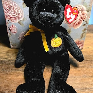 Può includere: Un orsacchiotto di peluche nero con una sottile finitura glitterata, caratterizzato da un nastro giallo legato al collo e una toppa ricamata a forma di zucca arancione sul petto. Un'etichetta rossa a forma di cuore Ty Beanie Babies è attaccata all'orecchio sinistro.