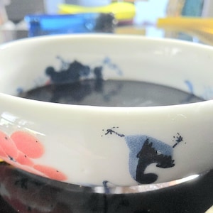May include: A white ceramic bowl with a dark liquid inside. The bowl is decorated with pink floral and blue abstract designs. The bowl sits on a reflective black surface.