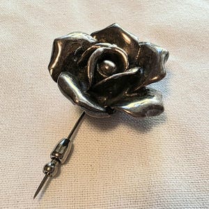 Può includere: Una spilla da bavero in metallo color argento con un design a fiore di rosa tridimensionale. Il fiore presenta petali curvi e stratificati con una finitura lucida. Un lungo e sottile stelo metallico si estende dalla base, accentuato da due piccole perline di metallo.