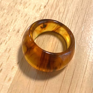 May include: A chunky, amber-colored ring with a marbled appearance. The ring has a rounded shape and a polished finish, showcasing a blend of warm brown and yellow hues. The ring is a statement piece of jewelry.