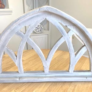 Op de afbeelding: Een verweerde, boogvormige raamkozijn in een lichtgrijze kleur. Het houten frame heeft een decoratief, opengewerkt ontwerp met elkaar kruisende gebogen en rechte lijnen. Het frame staat op een lichtbruine houten tafel.