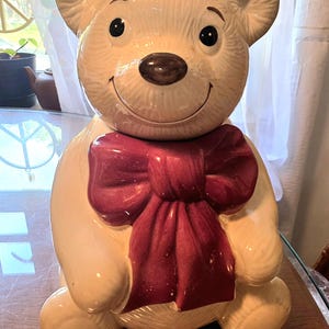 Puede incluir: Un tarro de galletas de cerámica con forma de oso sentado con un acabado brillante en color crema, ojos negros y nariz marrón oscuro. El oso presenta un lazo grande y texturizado de color rojo oscuro alrededor del cuello y detalles de pelaje.