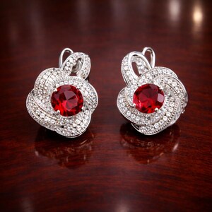 Peut inclure: Une paire de boucles d'oreilles argentées avec une pierre rouge centrale de taille ronde, entourée d'un motif en spirale superposé et incrusté de nombreuses petites pierres blanches claires qui captent la lumière avec éclat.