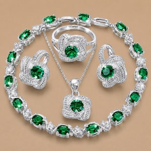 May include: A jewellery set consisting of a necklace, a ring, a pair of earrings, and a bracelet. Each piece features a central round-cut emerald green stone surrounded by a cluster of small, clear, sparkling white stones set in silver-toned metal.