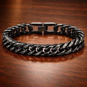 May include: A dark grey curb chain bracelet with a secure clasp. The bracelet features thick, interlocking links, creating a bold design. The metal has a matte finish, giving it a modern look. The bracelet is displayed on a wooden surface.