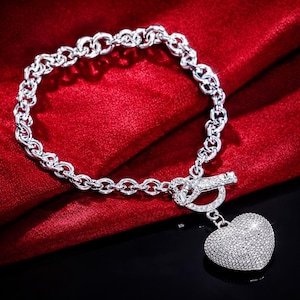 Peut inclure: Un bracelet argenté avec un motif de chaîne et un charm en forme de cœur. Le charm est orné de petites pierres scintillantes. Le bracelet est présenté sur un fond rouge.
