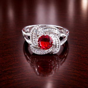 May include: A silver-toned ring featuring a large, round, faceted red centre stone. The stone is surrounded by a swirling, petal-like frame encrusted with numerous small, clear, sparkling white stones that extend along the band's shoulders.