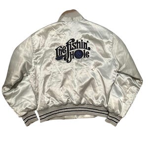 May include: A light silver satin bomber jacket with the text "The Fishin' Hole" embroidered on the back in navy blue. The jacket has a ribbed collar, cuffs, and hem with black and gray stripes. The jacket is a vintage style.