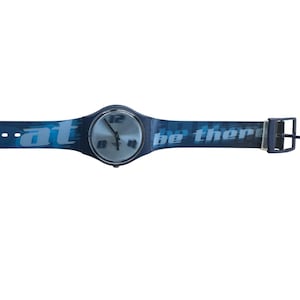 Puede incluir: Un reloj de pulsera azul con esfera plateada que muestra los números 12, 8 y 4. La correa es azul oscuro con el texto "at" en el lado izquierdo y "be there" en el lado derecho en color azul claro. El reloj tiene un cierre de hebilla estándar.