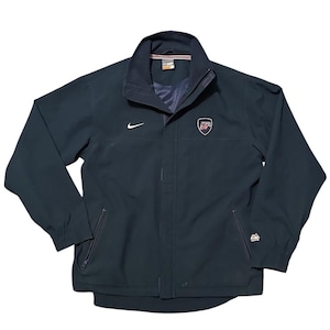 Könnte beinhalten: Marineblaue Jacke mit weißem Nike-Swoosh und USA-Emblem. Die Jacke hat einen durchgehenden Reißverschluss, zwei Vordertaschen und einen Stehkragen. Die Jacke ist ein leichter, sportlicher Stil.