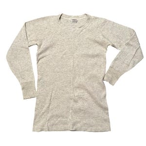 Pode incluir: Uma camisa térmica cinza claro de manga comprida. A camisa tem gola redonda e punhos canelados. O tecido tem uma aparência texturizada. A camisa está estendida sobre uma superfície branca.