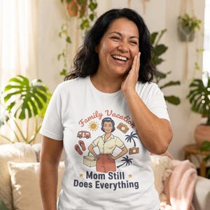 Könnte beinhalten: Weißes T-Shirt mit einem Aufdruck, der „Family Vacation“ und „Mom Still Does Everything“ lautet. Die Grafik zeigt eine Vintage-Illustration einer Frau, eines Wohnwagens, Kameras und Palmen. Das T-Shirt wird von einer Person getragen.