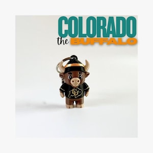 May include: A Colorado Buffaloes keychain featuring a cartoon bull mascot wearing a black and gold jersey and a black hat with gold trim. The text "COLORADO the BUFFALO" is above the mascot.