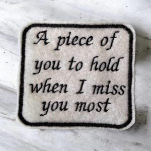 May include: A square-shaped off-white fabric patch with rounded corners and a thick black embroidered border. The patch features black embroidered text that reads: A piece of you to hold when I miss you most, written in a classic serif font style.