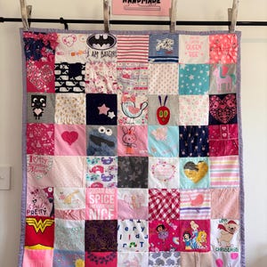 May include: A patchwork quilt made of various fabric squares featuring diverse prints, including cartoon characters, hearts, stars, and text like 'I AM BATGIRL', 'QUEEN BEE', 'SUGAR AND SPICE', and 'Lola'. The quilt has a light purple border and is hung by metal clips.