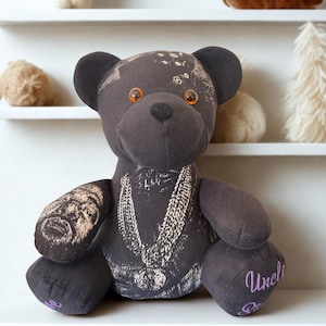 May include: A dark grey stuffed bear featuring a printed graphic of a person's face on the right arm, a chain necklace design on the chest, and orange plastic eyes. The word Uncle is embroidered in purple thread on the left foot of the bear.