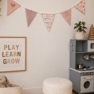 May include: A fabric bunting banner featuring five triangular pennants in shades of dusty pink and cream, decorated with small animal and floral illustrations, suspended from a white cord against a plain wall in a room with various toys.