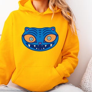 May include: A yellow hooded sweatshirt featuring a blue cat face graphic. The cat face has orange eyes, a pink nose, and a white-toothed grin. The sweatshirt has a front pocket and a drawstring hood.