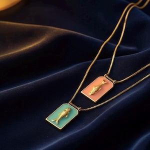 May include: Two gold-colored necklaces with rectangular pendants. Each pendant features a gold fish design on a colored background. One pendant is light pink, the other is light blue. The necklaces rest on a dark blue velvet surface.