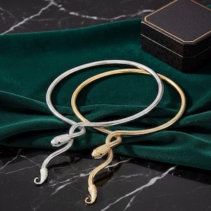 May include: Two snake-shaped necklaces. One is silver-toned, the other gold-toned. The snake heads are detailed with small embellishments. The necklaces rest on a dark green velvet surface, with a black jewelry box in the background.