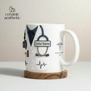 May include: White ceramic mug with a doctor's coat design, including a stethoscope, tie, and pocket details. Features the text "Enter Name" and a medical symbol. The mug sits on a wooden coaster.