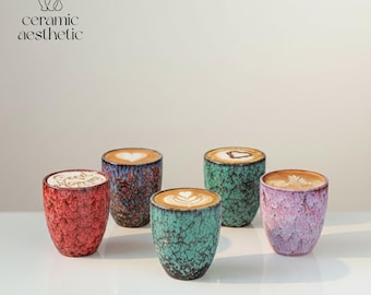 Può includere: Cinque tazze da caffè in ceramica con latte art, in rosso, blu, verde e viola. Le tazze hanno una superficie strutturata e sono piene di caffè. Il testo "ceramic aesthetic" è nell'angolo in alto a sinistra.