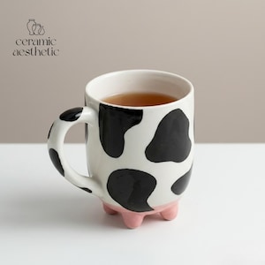 Peut inclure: Une tasse en céramique avec un motif de vache, présentant des taches noires sur un fond blanc. La tasse a une base rose en forme de sabots de vache et une anse incurvée. La tasse est remplie d'un liquide brun, et le texte "ceramic aesthetic" est visible.