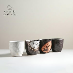 May include: Four ceramic cups in a row, each with a unique design. The first is white with a textured surface, the second is dark brown with white speckles, the third has a brown design, and the fourth is dark brown.