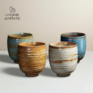 May include: Four ceramic tumblers in shades of blue, brown, and white are arranged on a white surface. The tumblers have a textured, handcrafted appearance, with visible brushstrokes and a rustic aesthetic. The text "ceramic aesthetic" is in the upper left corner.