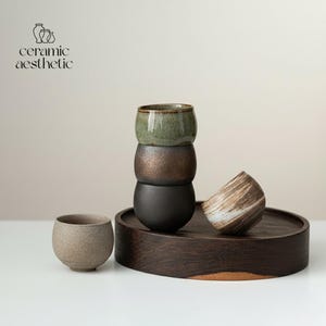 Può includere: Immagine di estetica ceramica con tazze rotonde in ceramica verde, bronzo e nera. Una tazza beige macchiata e un'altra con un motivo a spirale marrone e bianco, disposte su un vassoio di legno scuro.