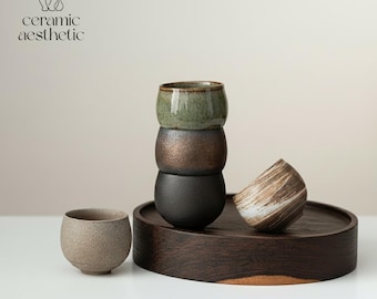 Peut inclure: Image de tasses en céramique. Une pile de trois tasses rondes, vertes, bronze et noires. Une tasse beige tachetée et une tasse à motifs tourbillonnants bruns et blancs sont également présentes, le tout sur un plateau en bois foncé.