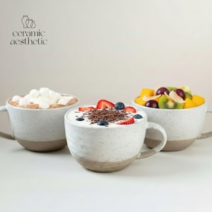 May include: Three speckled ceramic mugs with a light brown base. One mug contains hot chocolate with marshmallows, another yogurt with berries and chocolate shavings, and the third fruit salad. The text "ceramic aesthetic" is in the upper left corner.