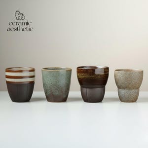 May include: Four ceramic cups in varying colors and designs. The cups are brown, green, and tan, with some featuring striped or speckled patterns. The text "ceramic aesthetic" is in the upper left corner.