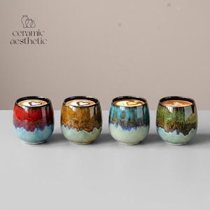 May include: Four ceramic coffee cups with a unique, colourful glaze. Each cup has a different colour scheme, including red, brown, teal, and green, with a light blue base. The cups are filled with coffee and topped with a heart-shaped design.