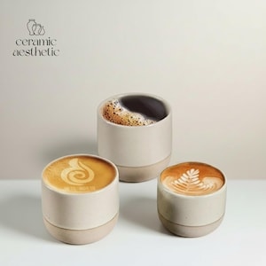 Op de afbeelding: Drie keramische koffiekopjes met latte art en een kopje zwarte koffie. De kopjes zijn gebroken wit met een bruine basis. De tekst "ceramic aesthetic" staat linksboven.