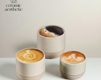 Peut inclure: Trois tasses à café en céramique avec latte art et une tasse de café noir. Les tasses sont blanc cassé avec une base beige. Le texte "ceramic aesthetic" est en haut à gauche.