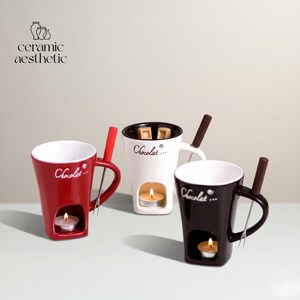 May include: Three ceramic fondue mugs in red, white, and dark brown, each featuring the word Chocolat printed on the front. Each mug includes a built-in base compartment for a tea light candle and a matching fork resting in the handle.