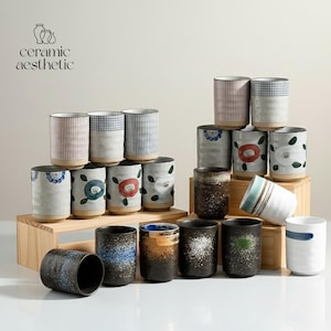 May include: A selection of ceramic mugs with diverse patterns, including floral, striped, and speckled designs. The colour scheme features white, blue, red, and brown. The mugs are displayed on a wooden stand. The text "ceramic aesthetic" is visible.