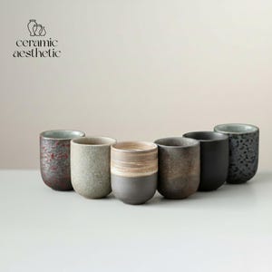May include: Seven ceramic cups of varying colours and textures are displayed. The cups are cylindrical with a subtle taper. Colours include speckled red, grey, and solid black. The text "ceramic aesthetic" is visible in the top left corner.