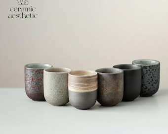Peut inclure: Collection de sept tasses en céramique de différentes couleurs et textures. Les tasses sont cylindriques avec une forme légèrement conique. Les couleurs varient du rouge foncé tacheté au noir uni. Le texte "ceramic aesthetic" est visible.