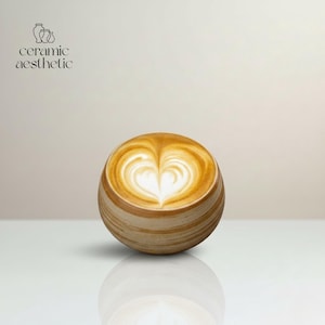 Puede incluir: Una taza de café cerámica esférica con un diseño de arte latte en forma de corazón. La taza tiene un exterior marrón claro a rayas y está llena de una bebida cremosa de color marrón claro. El texto "ceramic aesthetic" está en la esquina superior izquierda.