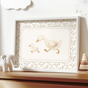 Pode incluir: Uma impress&atilde;o emoldurada com uma ilustra&ccedil;&atilde;o em aquarela de uma m&atilde;e pata e seu patinho. A impress&atilde;o tem uma moldura branca e uma borda floral. Uma figura de elefante branco e um brinquedo de empilhamento de madeira est&atilde;o na superf&iacute;cie de madeira.