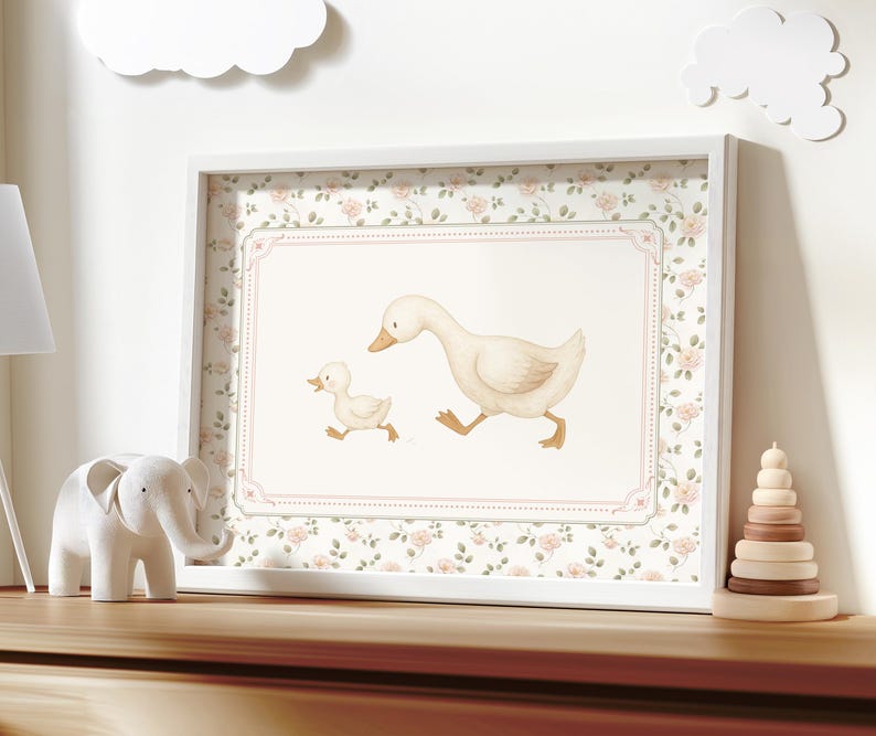 Pode incluir: Uma impress&atilde;o emoldurada com uma ilustra&ccedil;&atilde;o em aquarela de uma m&atilde;e pata e seu patinho. A impress&atilde;o tem uma moldura branca e uma borda floral. Uma figura de elefante branco e um brinquedo de empilhamento de madeira est&atilde;o na superf&iacute;cie de madeira.