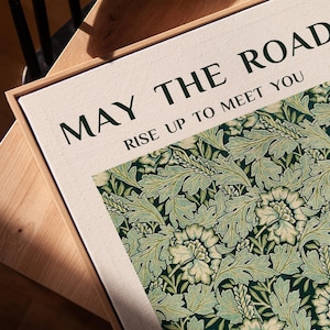 Może przedstawiać: Oprawiony wydruk na pł&oacute;tnie z napisem "MAY THE ROAD RISE UP TO MEET YOU" i kwiatowym wzorem w odcieniach zieleni i kremu. Wydruk ma ramę z naturalnego drewna i jest umieszczony na drewnianej powierzchni.