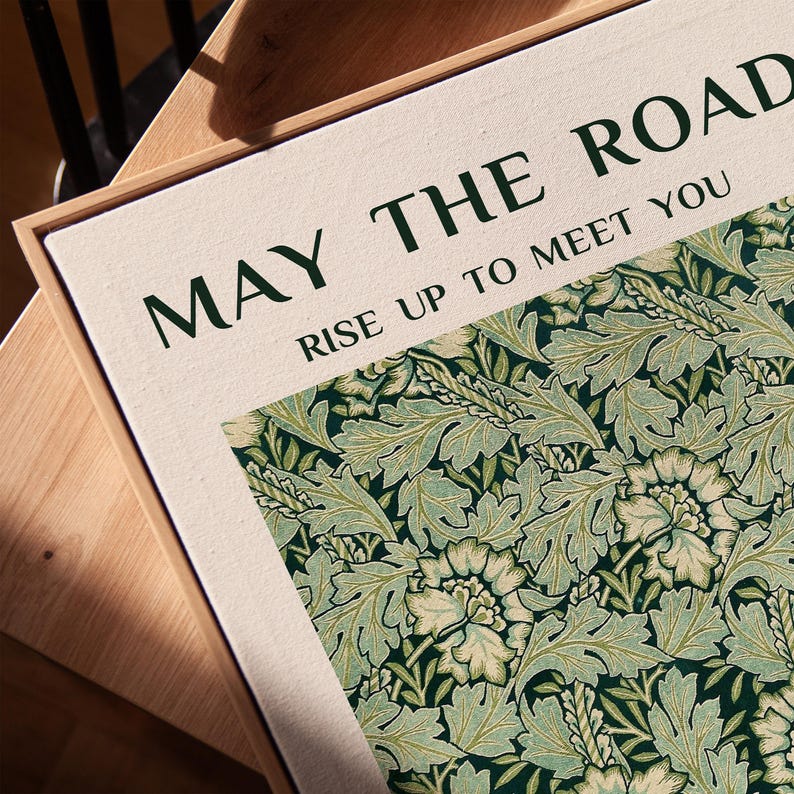 Może przedstawiać: Oprawiony wydruk na pł&oacute;tnie z napisem "MAY THE ROAD RISE UP TO MEET YOU" i kwiatowym wzorem w odcieniach zieleni i kremu. Wydruk ma ramę z naturalnego drewna i jest umieszczony na drewnianej powierzchni.