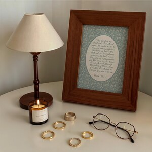 May include: A lit candle in a glass jar, a wooden framed print with text, and several gold rings are arranged on a white surface. A small lamp with a beige shade and round eyeglasses complete the scene. The framed print contains the text of Psalm 23:1-4.